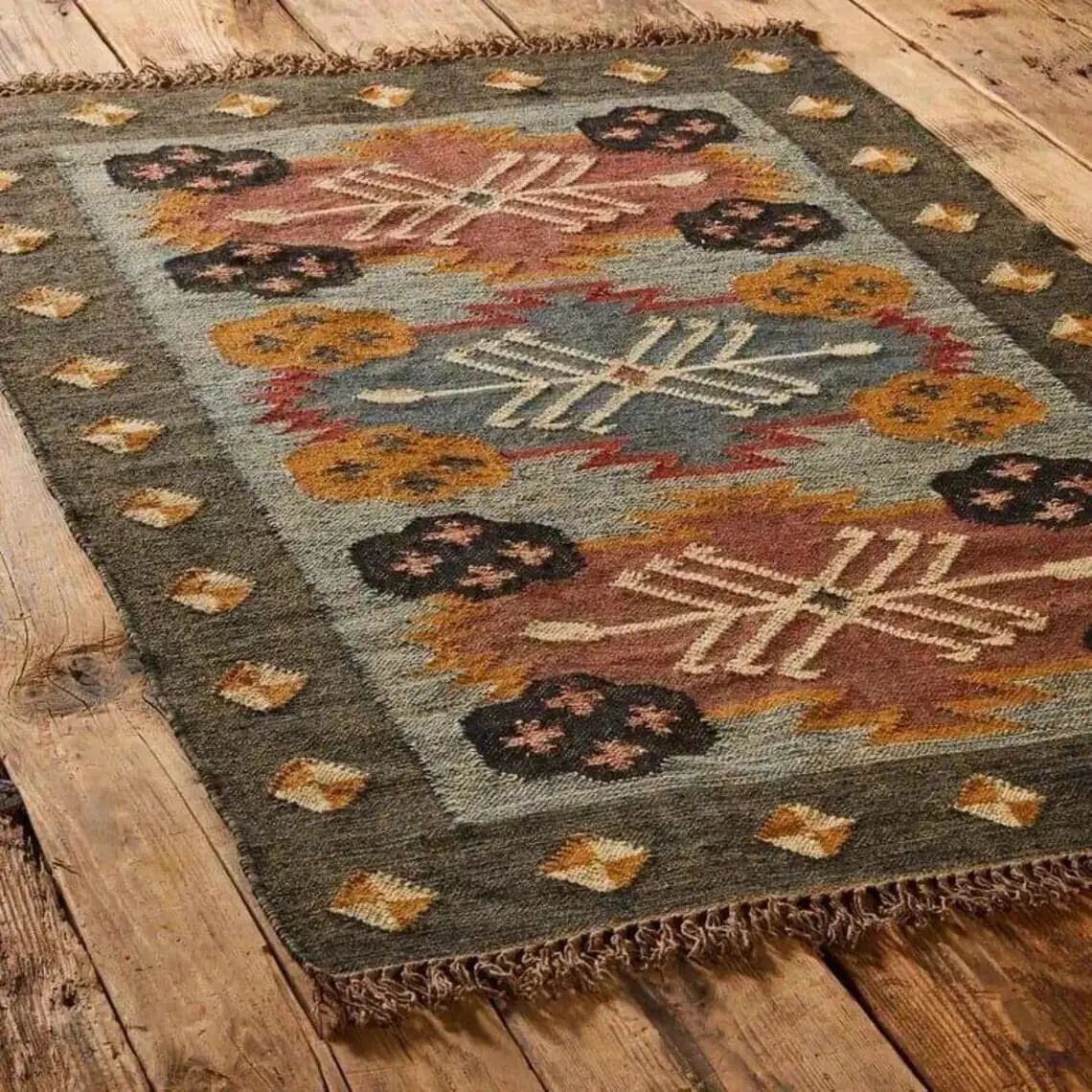 Flat-woven rug on a wooden floor with an olive border and cream starburst medallions on rose and grey-blue panels